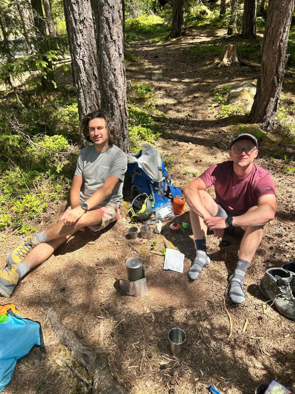 Dominik und Thomas sitzen bei einer Bergtour im Wald, dazwischen ein Kocher und zwei Becher